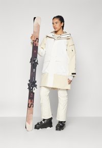 Ski jacket in white and beige, featuring a hood and multiple pockets. The model holds brown skis with black bindings. Wearing black ski boots.
