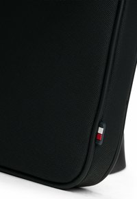 Borsa in tessuto nero textured con bordi arrotondati, caratterizzata da un piccolo logo rosso, bianco e blu sul lato. Rifiniture lisce e design robusto.