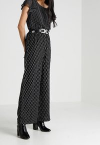 Combinaison noire à pois avec manches volantées, cintrée à la taille par une ceinture argentée. Dotée d'un design à jambes larges et de talons noirs brillants.