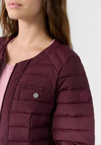 Bordeaux puffer jack met ritssluiting, horizontale gestikte textuur en een borstzak met een ronde drukknoop. Logo op de mouw.