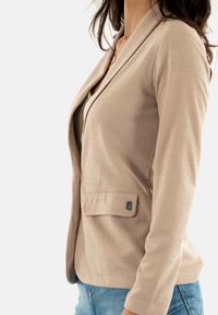 Blazer beige con tessuto testurizzato, design aderente, colletto a scialle e tasca laterale singola con dettaglio metallico. Indossato sopra una blusa chiara.