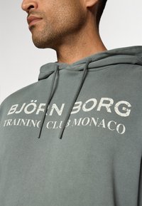 Zöld pamut keverék kapucnis pulóver elülső grafikával: "BJÖRN BORG TRAINING CLUB MONACO" elhasználódott fehér betűtípussal.