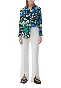 Langarmshirt met een kleurrijk abstract patroon in blauw, groen, zwart en wit, gecombineerd met witte flarebroeken en zwarte sandalen.