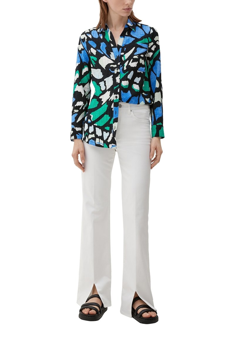 Langarmshirt met een kleurrijk abstract patroon in blauw, groen, zwart en wit, gecombineerd met witte flarebroeken en zwarte sandalen.