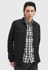 Schwarze Button-Up-Hemdjacke mit zwei Brusttaschen, über einem schwarz-weißen karierten Hemd getragen. Strukturierter Stoff mit passgenauem Schnitt.