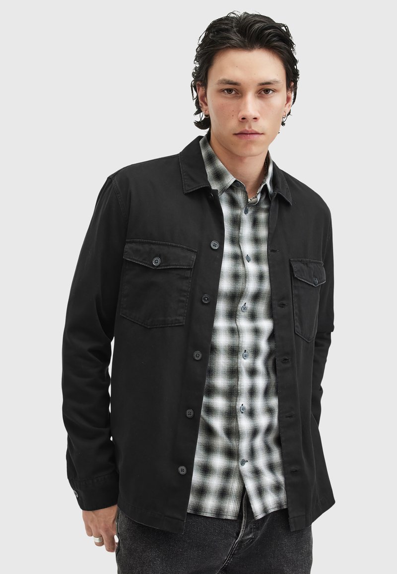 Schwarze Button-Up-Hemdjacke mit zwei Brusttaschen, über einem schwarz-weißen karierten Hemd getragen. Strukturierter Stoff mit passgenauem Schnitt.