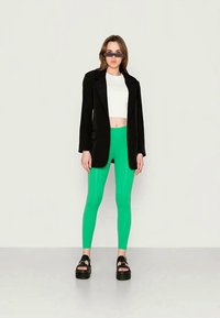 Leggings verdes feitos de material elástico, combinados com um top cropped branco e um blazer preto. A modelo usa óculos de sol oversized e sandálias de plataforma.