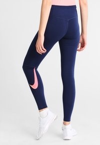 Kvinna i marinblå leggings med en rosa swoosh-logga på vänster vad och vita sportskor, står mot en enfärgad bakgrund.