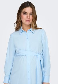 Γυναίκα με καστανά μαλλιά μήκους μέχρι τους ώμου ς, που φοράει ανοιχτό μπλε φόρεμα με κουμπιά και υφασμάτινη ζώνη δεμένη στη μέση.