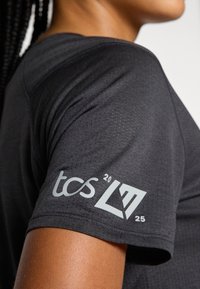 Camiseta atlética negra con tejido texturizado, mangas cortas y un logo impreso en blanco en el brazo izquierdo con acentos geométricos.