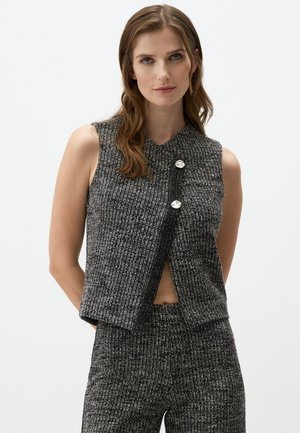 Ärmelloses schwarz-weißes Cropped-Top mit geripptem Muster, das einen V-Ausschnitt aufweist und zwei dekorative silberne Knöpfe auf der Vorderseite hat.