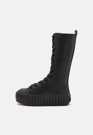 Vysoká černá šněrovací bota s hrubou vroubkovanou podrážkou a poutkem na patě, zobrazena z boku na jednobarevném pozadí.