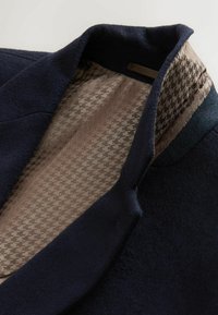 Marinblå ullkappa med krage, som visar en texturerad beige foder med houndstooth-mönster. Inre etikett synlig nära kragen.