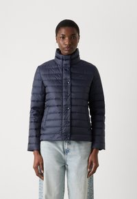 Giubbotto imbottito blu navy con colletto alto, chiusura a bottoni, design trapuntato e maniche lunghe. Indossato sopra jeans azzurri chiari.
