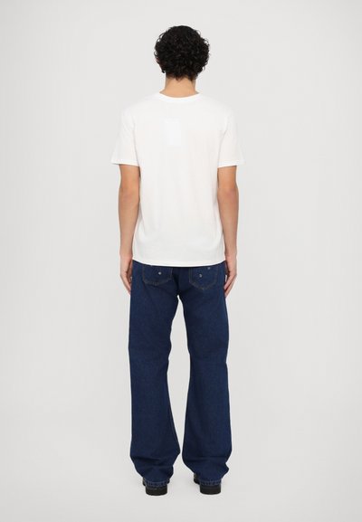 Weißes T-Shirt mit kurzen Ärmeln, kombiniert mit blauen weit geschnittenen Jeans. Das T-Shirt hat einen schlichten Rücken; die Jeans verfügen über einen Knopfverschluss und Gesäßtaschen.