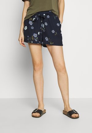 Kvinde iført marineblå blomstrede shorts med bindebånd i taljen, olivengrøn top og sorte klipklappere med store spænder.