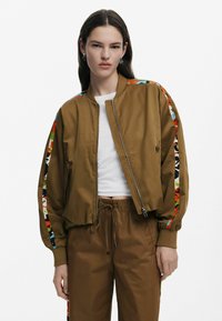 Hnedá letecká bunda so zipsom vpredu, rebrovanými manžetami a farebnými vzorovanými akcentmi na rukávoch. Tkanina vyzerá hladko a mierne leskne.