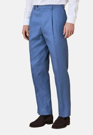 Man wearing tailored light blue dress pants with front pleats, paired with a white shirt and dark brown loafers, standing against a plain background.