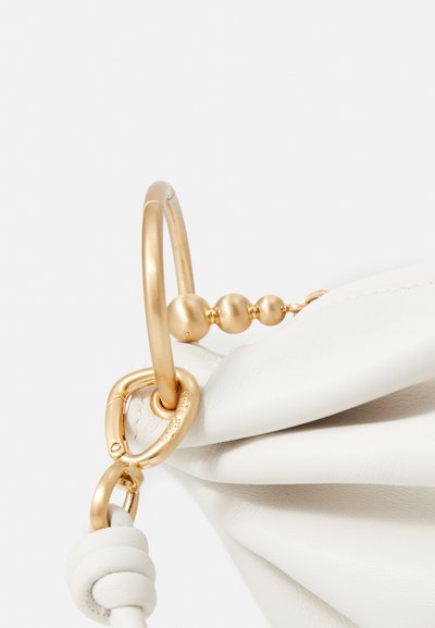 Balta ādas rokassoma ar zeltainu gredzena rokturi un sfēriskām akcentiem, prezentējot mīkstu, plīsušo tekstūru.