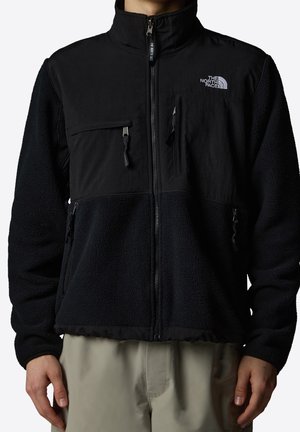 Czarna kurtka polarowa z zamkiem błyskawicznym, kieszeniami na klatce piersiowej i po bokach zamykanymi na zamek oraz białym logo The North Face na górnej części klatki piersiowej.