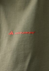 Olivově zelené tričko z tkaniny s výrazným červeným logem "H.KOUMORI" a malým červeným detailem trojúhelníku Adidas, hladká textura.
