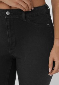 Jeans de mezclilla negra con cintura alta, bolsillos frontales, un botón de metal y una tela suave y texturizada. Vista cercana de la cintura.