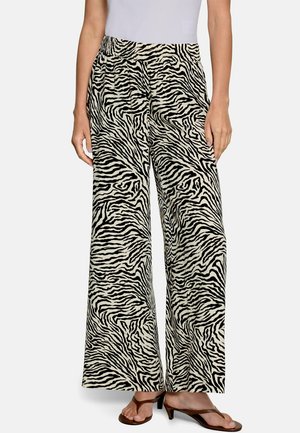 Femme portant un pantalon large avec imprimé zèbre noir et blanc et des sandales marron à talons, debout devant un fond uni.