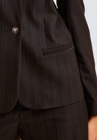 Zwarte pinstripe blazer met een gestructureerde pasvorm, voorzien van een enkele metalen knop en een paspelzak aan de voorkant. Gertexteerde stof met fijne lijnen.
