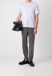 Mężczyzna w jasnofioletowej koszulce, szarych dżinsach, czarnych mokasynach z białymi skarpetami, trzymający dużą czarną skórzaną kopertówkę.