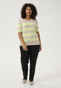 Pulover scurt cu mânecă, cu dungi bej și verde neon, asortat cu pantaloni negri și pantofi de culoare deschisă, stând pe un fundal simplu.
