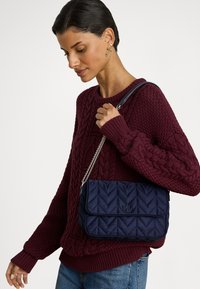 Μπλε καπιτονέ τσάντα με αλυσίδα, σχεδιασμένη με μοτίβο ψαροκόκαλου και ανάγλυφο λογότυπο. Συνδυασμένη με πουλόβερ μπορντό με πλέξη καλώδιο.