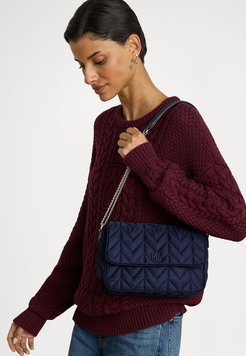 Μπλε καπιτονέ τσάντα με αλυσίδα, σχεδιασμένη με μοτίβο ψαροκόκαλου και ανάγλυφο λογότυπο. Συνδυασμένη με πουλόβερ μπορντό με πλέξη καλώδιο.