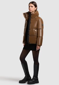 Jachetă puf cu un ton de maro, cu guler înalt, material lucios, nasturi cu presiune și buzunare laterale; purtată cu o rochie neagră cu textură rib și cizme negre până la genunchi.