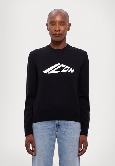Suéter negro de punto con escote redondo, con un logo blanco en el pecho. Combinado con jeans de mezclilla azul claro.