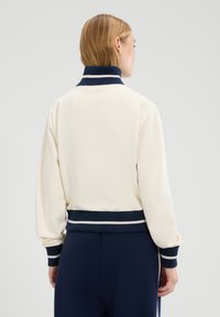 Chaqueta color crema con un cuello y puños de punto en navy, con una franja blanca en el cuello y la cintura. Tejido suave, corte relajado.
