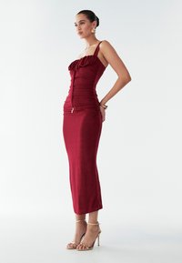 Burgundská midi šaty s volánovým výstrihom, priliehavým telom a hladkou textúrou. V kombinácii s páskovými zlatými sandálmi na vysokom podpätku.