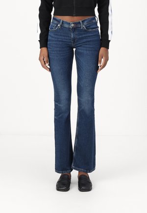 Woman wearing dark blue flared jeans, black cropped jacket, and black loafers standing against a white background.