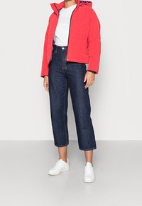 Rød quiltet jakke med lynlås og hætte, parret med højdelte mørkeblå jeans og hvide sneakers. Jakken har et mønstret indre foder.