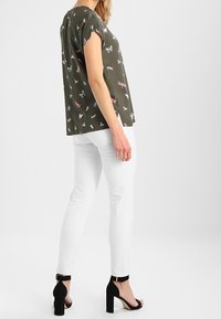 Frau in einem olivgrünen Shirt mit buntem Insektenmuster, weißen Hosen und schwarzen High Heels, die seitlich steht.