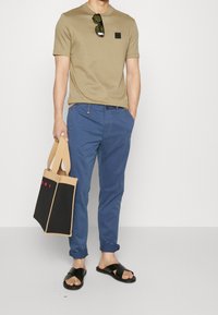 T-shirt à manches courtes beige avec écusson logo, pantalon bleu, sandales noires à enfiler, et sac fourre-tout beige et noir avec lettrage rouge.