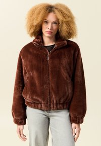 Brązowa, pluszowa kurtka bomberka na zamek z wysokim kołnierzem i elastycznymi mankietami, zestawiona z jasnoszarymi dżinsami.