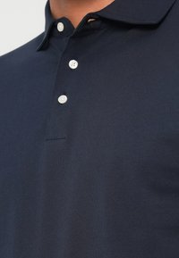 Tumši zils polo krekls ar mīkstu, strukturētu audumu, ar apkakli, trim baltām pogām un smalku pogcaurumu detaļu.