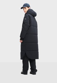 Dlhý čierny páperový kabát s kapucňou, s prešívaným dizajnom a bočnými rozšireniami pre ľahký pohyb. Kombinovaný s tmavými nohavicami a zodpovedajúcou čiapkou.