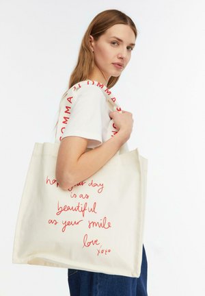 Młoda kobieta niosąca kremową torbę na ramię z czerwonym, ręcznie pisanym napisem i markowymi paskami na ramiona, ubraną w białą koszulkę i dżinsy.