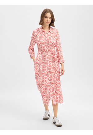 Jonge vrouw draagt een midi-jurk met roze-wit patroon en een riem, loopt vooruit en draagt witte sneakers tegen een effen achtergrond.