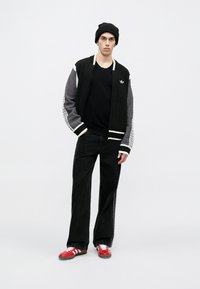 Μαύρο πλεκτό μπουφάν τύπου bomber με γκρι μανίκια, μαύρο T-shirt, φαρδιά μαύρη τζιν παντελόνια και κόκκινα αθλητικά παπούτσια με λευκές ρίγες. Μαύρος σκούφος.