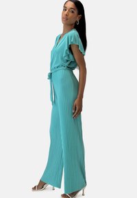 Turkos plisserad jumpsuit med korta volangärmar, V-ringning och ett tygbälte. Slät textur och vida ben.