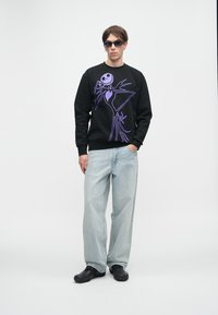 Czarna bluza z nadrukiem fioletowego szkieletu, jasnoniebieskie luźne dżinsy i czarne buty. Model nosi okulary przeciwsłoneczne i stoi na białym tle.