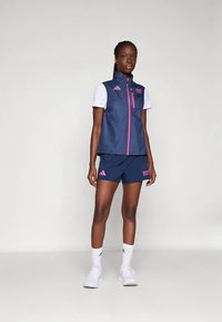 Tengerészkék sportmellény rózsaszín cipzárral, fehér pólóval és rózsaszín díszítéssel ellátott tengerészkék rövidnadrággal párosítva. Könnyű anyagból készült.