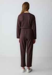Bruine jumpsuit met lange mouwen, comfortabele pasvorm, elastische tailleband en taps toelopende pijpen, gecombineerd met witte sneakers. Soepele stoftextuur.